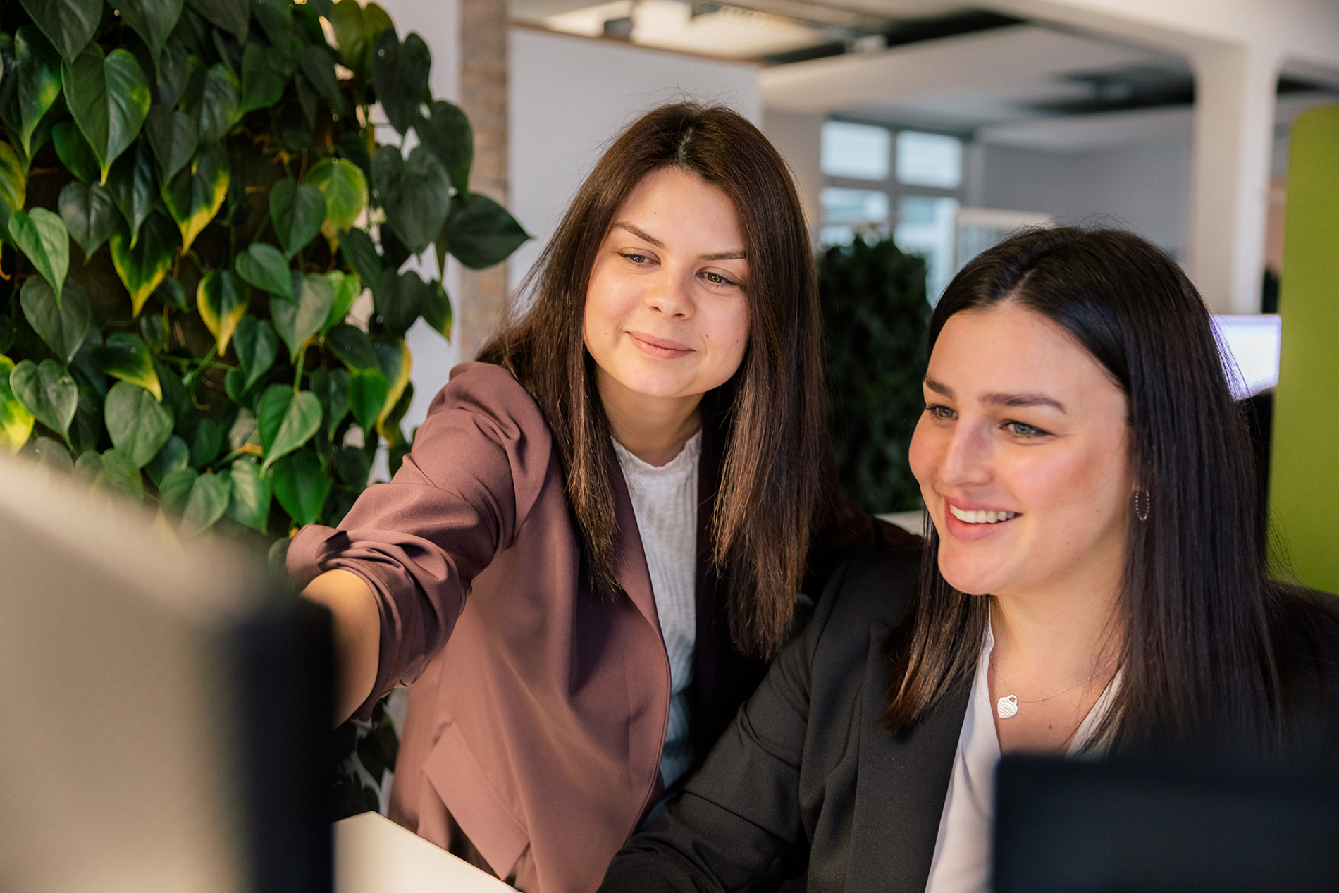 Zwei Frauen arbeiten an einem modernen Bürotisch, umgeben von üppigen Grünpflanzen, konzentriert auf einen Computerbildschirm.